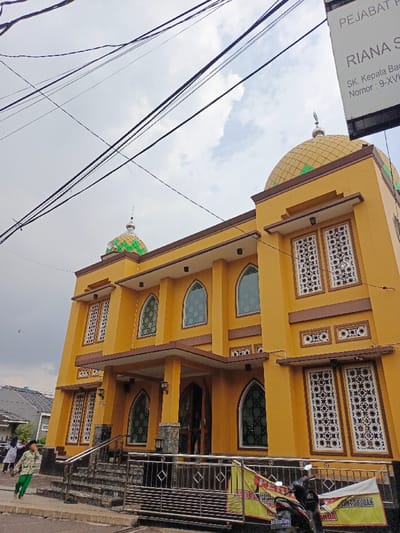A yellow, two-story building with a gold dome stands prominently. The structure has several windows with decorative white screens. Another smaller dome, colored green, is also visible. Power lines crisscross the cloudy sky above. A small flight of stairs leads up to a covered entrance. A motorcycle and a banner partially obscure a metal railing in front. People are walking on the sidewalk. A sign is visible on the building's edge.