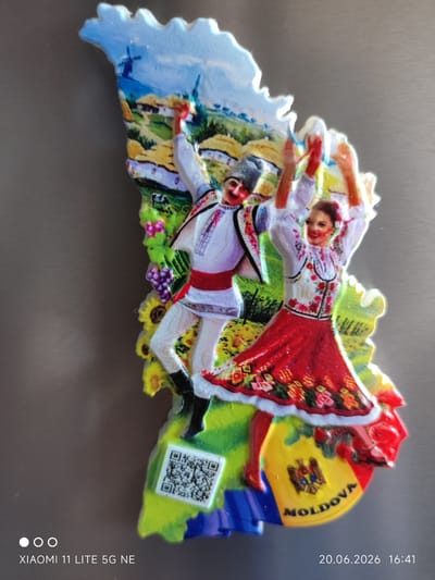 The image is a close-up of a souvenir magnet shaped like the country of Moldova, featuring a depiction of two dancers in traditional Moldovan attire. The dancers are in mid-motion, with one male dancer on the left and one female dancer on the right. The male dancer wears a white shirt with red and black trim, a red sash, and a fur hat. The female dancer wears a white embroidered blouse with a red skirt adorned with floral patterns. Behind the dancers, the magnet shows a rural landscape with houses, fields, a windmill, sunflowers, and grapes. The national emblem of Moldova is visible on a yellow and red background at the bottom of the magnet, with the word "MOLDOVA" written in black. A QR code is also present on the magnet. The magnet is placed on a metallic, possibly stainless steel surface, which appears to be a refrigerator door. The timestamp "20.06.2026 16:41" and "XIAOMI 11 LITE 5G NE" are visible at the bottom of the image, suggesting it was taken with a Xiaomi phone on that date and time. The location of Böblingen, Germany is not visually represented in the image, nor is it directly inferable from the content of the souvenir magnet itself.