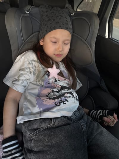 A young girl is captured sleeping peacefully in a car seat within a vehicle. She is positioned on her right side, eyes closed, suggesting deep rest or sleep. She wears a grey beanie with a subtle leopard print, a light-colored t-shirt featuring graphic designs in black and purple, and dark wash jeans. A small, pink, star-shaped embellishment is visible on her t-shirt near her shoulder. Her right hand, encased in a black and white striped fingerless glove, holds a small, light-brown, textured object. The car's safety belt is buckled across her chest. The interior of the car appears somewhat dark, with natural light entering from a window on the left, indicating daytime. Visible text on her t-shirt includes the word "SUPER" and partially visible words like "AWESOME" and "EVERY DAY". The scene portrays a tranquil moment during travel, set in what is identified as Yus'ki, Russia.
