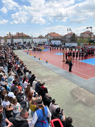 The image captures a cultural event or performance taking place outdoors in the city of Rakovski, Bulgaria. A large crowd of people is seated on tiered benches, facing a central red and blue sports court or stage area. Performers dressed in traditional black and red costumes, possibly participating in a folk dance or parade, are arranged on the court. A wooden cross is being carried by a person in a black jacket with a patterned cloth. Banners and signs with text "ORIGAMI", "КАЛИМАНА", "Emjaya", "Via Déco", "Олдинг", "grips pvc", "ЖИВ ПЛЕТ", "Arios pro", "ВАЛИКДЕНСКИ" and "JERN HA" are visible near the performers. Buildings with red-tiled roofs are visible in the background, and a partly cloudy blue sky suggests daytime.