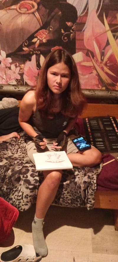 A young female is sitting on a bed in what appears to be a bedroom. She has long, brown hair and is wearing a gray tank top and light-colored shorts. She is holding a pen and appears to be drawing in a sketchbook that is on her lap. A smartphone is visible on her right leg, displaying an image. The background shows a tapestry with floral and ornamental designs. The floor is visible in the lower portion of the image, and a white sandal is on the floor near her foot. The lighting suggests an indoor setting, possibly during the evening. No specific location in Chernyavskii, Russia can be determined from the visual information provided in the image.
