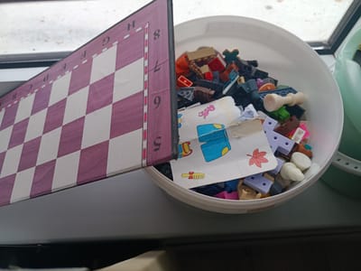 A white plastic tub filled with assorted colorful Lego-like building blocks and other small toys sits on a windowsill in Arkadag, Turkmenistan. A portion of a purple and white checkered chessboard, with letters and numbers printed along its edge, is propped up against the tub, partially obscuring the toys. Some of the visible toy pieces include red, blue, tan, brown, gray, and pink blocks, as well as a white chess piece resembling a pawn. A piece of paper with cartoon drawings of a pink dragon and a red leaf is also visible among the toys. The scene is viewed from above and to the side, with the background showing a window with a view of an outdoor environment.