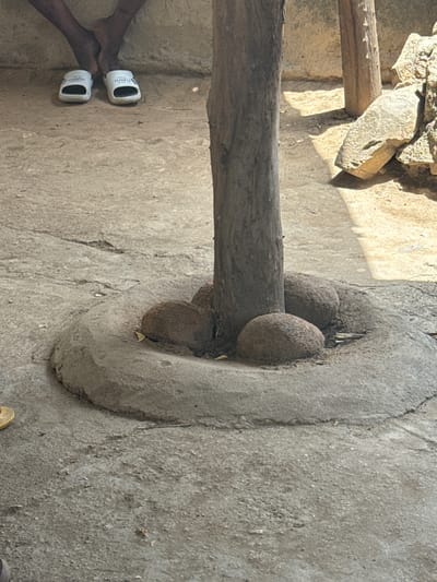 The image, taken in Dawaki, Nigeria, shows a weathered, cylindrical, stone or concrete structure embedded in a rough, gray concrete or dirt ground. A thick, gray tree limb passes horizontally through a central opening in the structure, supported by three spherical, textured objects resting inside the opening against the limb. Nearby, a pair of dark-skinned feet wearing white sandals are visible. Fragments of rock and another rough wooden beam are partially visible in the upper left corner of the frame.