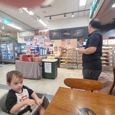 The image depicts an indoor scene, likely a bakery or a section of a supermarket, with a focus on baked goods. In the foreground, a young child is seated in a high chair, looking to the left. The child is wearing a black and white shirt with "Aloha! Beach" printed on it. To the right of the child, a wooden table and chair are visible, suggesting a dining or waiting area.

In the middle ground, a man in a dark blue polo shirt and grey shorts is standing and looking to the left. Behind him, a display of various baked goods is arranged on shelves. Stacks of red pizza boxes are also prominent. A green bin with signage, possibly for waste disposal, is located to the left of the baked goods display.

The background reveals more of the store environment, including refrigerated display cases containing food items, promotional posters with images of pizza and baked goods, and overhead lighting fixtures. A sign with Korean text, possibly indicating prices or product names, is visible above the pizza boxes. The overall lighting is bright and consistent with an indoor commercial establishment. The presence of a sign with "New York Pizza" suggests a possible affiliation with American-style pizza. The image appears to be taken at a "New York Pizza" location within a larger store in Wonju-si, South Korea.