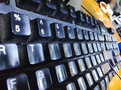 The image is a close-up, high-angle shot of a black computer keyboard. The keys are worn, with some showing signs of dust and grime. The focus is on the keys themselves, with the background blurred and showing a yellow surface and a hint of something wrapped in yellow material with a bow. The keyboard appears to be a standard QWERTY layout with some function keys visible at the top. The keys show alphanumeric characters and symbols. The overall impression is of a used keyboard. There is no visible indication of the city of Rumuepirikom, Nigeria, in this image; it solely depicts a keyboard.