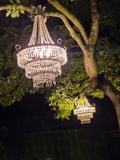 An ornate, multi-tiered crystal chandelier hangs from a tree branch at night, illuminated by its own lights. The chandelier features several rows of dangling crystals that reflect the warm light from within. A second, smaller chandelier of similar design is visible in the background, also hanging from a tree. Lush green foliage surrounds the chandeliers, and the dark night sky serves as a backdrop. The scene is set outdoors, likely in a garden or park. The location is specified as being in the city of Bettembourg, Luxembourg, though no specific landmarks or distinguishing features of the city are visible in the image. There are no people or animals present.