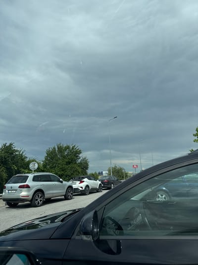 The image captures a daytime scene from within a dark-colored vehicle, likely parked, looking out over a parking lot or roadside area. The foreground on the right is dominated by the interior of the car, showing the steering wheel, dashboard, and the side mirror.
A line of cars is parked facing away from the viewer on the left side of the frame. The most prominent is a silver Volkswagen Tiguan SUV, clearly visible in the mid-ground. Its rear license plate, "CT 6873 PT," indicates a Bulgarian registration (Stara Zagora region), aligning with the provided location context of Radinovo, Bulgaria. Behind the silver SUV, a white SUV and another dark car are also parked.
The setting includes scattered green trees behind the parked vehicles and tall streetlights or lamp posts extending into the distance. A circular convex mirror on a pole is visible near the silver SUV. In the far background, a red and white sign, possibly for a gas station or shop, can be seen, though the text is indistinct due to distance. A white truck with a blue logo is partially visible beyond the sign.
The sky is partly cloudy and bright, suggesting a clear day with some atmospheric haze or wispy clouds. The overall impression is that of a stop in a commercial or transit area. No people are visible, and no specific event or activity other than parking is taking place.
