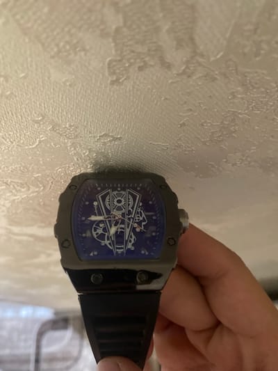 The image shows a close-up of a person's hand holding a watch with a dark gray casing and a dark blue dial. The watch face features a complex, mechanical design with gears and hands, rendered in a light color against the blue background. Visible text on the dial appears to be "Shangfuci". The watch is attached to a black strap. The background is a textured, light-colored surface, possibly wallpaper, with some areas of shadow and light suggesting indoor lighting. The overall lighting is somewhat dim. The context provided indicates the location is Istaravshan, Tajikistan, but there are no overt visual cues within the image to confirm this specific location. The image focuses on the watch and the hand holding it, with no other subjects or clear environmental details visible.