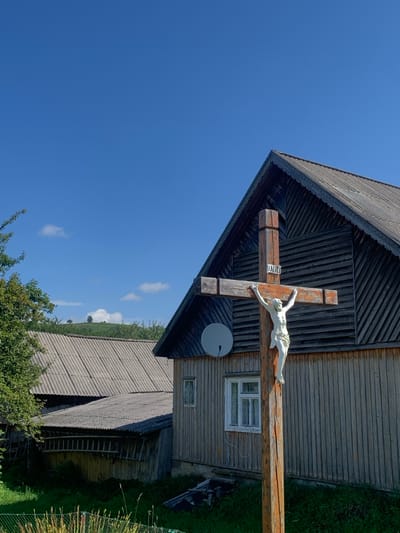 This image, taken in Lazeshchyna, Ukraine, displays a large, prominent wooden crucifix in the foreground, positioned slightly right of center. The crucifix is light brown, featuring a white, three-dimensional figure of Christ and the inscription "INRI" near the top of its vertical beam. Directly behind the crucifix, to the right, stands a two-story house constructed of dark, vertical wooden planks with a steeply pitched grey corrugated roof that includes decorative trim along the eaves. A white-framed window and a circular grey satellite dish are visible on the house's facade. To the left and set back, a second building with a grey corrugated roof is partially visible, featuring a lower, darker section. The immediate foreground shows green grass and low vegetation. The background features a clear blue sky with sparse white cumulus clouds, above which green, tree-covered hills are visible on the horizon. The scene is illuminated by direct sunlight, indicating daytime conditions.