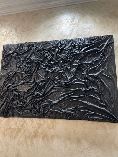 A photograph depicts a rectangular black sculpture, seemingly made from tightly wrinkled plastic sheeting, mounted on a beige textured wall. The sculpture is positioned vertically, taking up a significant portion of the frame, with the wall visible on either side. The left side of the frame shows a white door frame. The photograph may have been taken indoors in Лесной Пейзаж Кп (шарапово), Russia. There are no people visible in the photo.