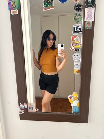 A person in sunglasses, a mustard-colored short-sleeved top, and black shorts takes a mirror selfie. The mirror has a brown frame covered with stickers. The stickers include cartoon characters like Homer Simpson, Courage the Cowardly Dog, and other pop culture images. The background shows a white wall and a glimpse of a wooden floor. The person holds a white smartphone.
