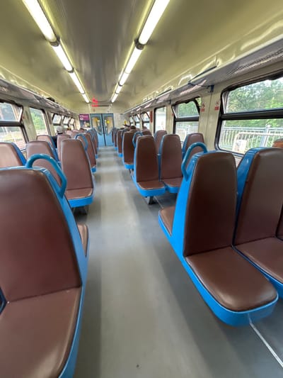 The image shows the interior of a train carriage, likely a commuter train or "elektrichka," consistent with the Komarovo, Russia location context. The perspective is tilted 90 degrees, looking down a long, central aisle between two rows of seats.
The seating features warm brown upholstered cushions set within distinct bright blue frames and armrests. The seats are arranged in pairs, facing each other across the aisle. The floor of the carriage is a utilitarian gray, contrasting with the light beige or off-white ceiling, which is fitted with long, bright fluorescent or LED tube lights running the length of the carriage.
Large windows line both sides, revealing green trees and some distant structures, indicating daylight and a journey through a natural or suburban setting. Blue grab handles or horizontal bars are visible above the windows on the left side, likely for standing passengers.
Towards the far end of the carriage on the left, a single passenger is partially visible, seated. In the distance, a red digital display shows the numbers "20 12," which could indicate the time or a route number. Above this display, the number "60" is faintly visible. Small, red pictogram signs are also present above the windows on the right side. The carriage appears clean and mostly empty, suggesting it is not at peak occupancy.