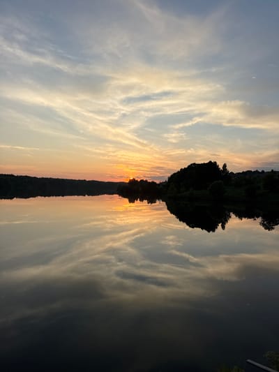 A wide shot captures a tranquil lake at dusk. The sky is a canvas of soft blues and yellows, with wispy clouds reflecting on the still water. The sun, partially obscured by trees on the horizon, casts a warm glow. Dense foliage lines the far bank of the lake, creating a dark silhouette against the vibrant sky. A few scattered houses are visible nestled within the trees. The water is dark and reflects the sky and landscape with remarkable clarity, creating a mirror image of the scene above. The overall atmosphere is one of peaceful serenity. The location is identified as the city of Kamysh, Russia.