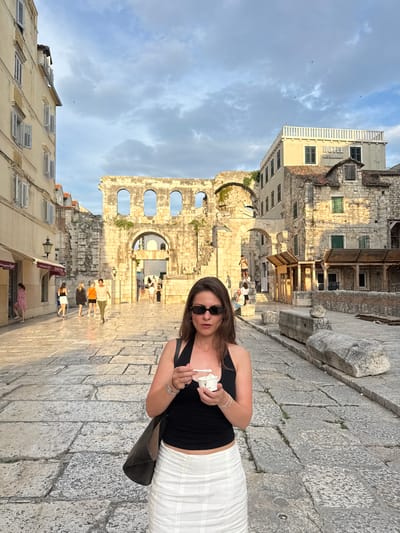 The image shows a young woman in a black tank top and white skirt, wearing sunglasses, holding a small white cup. She is standing on a cobblestone street in front of ancient stone ruins with arched windows. The ruins are made of large, weathered stone blocks, some of which are crumbling. Sunlight casts shadows on the stone, suggesting it is either late afternoon or early morning. In the background, several other people can be seen walking through the ruins and along the street. To the left of the ruins, a modern building with a white railing and windows is visible. The sky is blue with scattered clouds. The overall scene appears to be a tourist area in a historic city. The text "HOTEL" and "RESTAURANT" is visible on the modern building in the background.