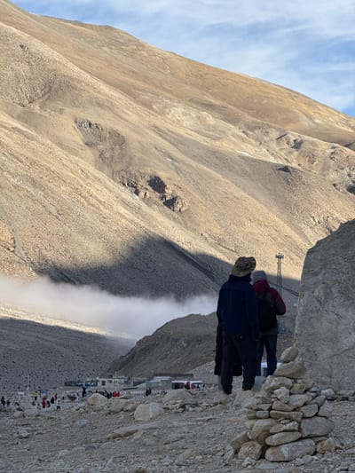 The image depicts a mountainous landscape near Tashi Dzom town, China. The scene includes people standing on a rocky outcrop, with a steep, sparsely vegetated mountainside in the background. In the distance, a building complex is visible. A plume of dust or mist rises in the foreground.