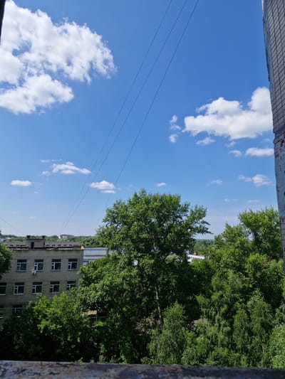 The image depicts an outdoor scene, likely from a high vantage point such as a window or balcony, overlooking a residential area with lush greenery. The sky is a vibrant blue, dotted with scattered white clouds, suggesting a clear and pleasant day. Overhead power lines are visible stretching across the frame, indicating an urban or suburban setting.
In the foreground, to the left, the edge of a building or structure with a tiled roof is partially visible. Dominating the right side of the image is a dense canopy of green trees, their leaves creating a rich texture.
Beneath the trees and partially obscured by them, a multi-story brick building with numerous windows is evident. The building appears to be residential, with a somewhat utilitarian architectural style. The overall impression is one of a tranquil, possibly summer day in a Russian locale like Ерино, where nature and urban development coexist. The time of day appears to be daytime, given the bright sky and shadows. No people are clearly discernible in the image, and there is no visible text. The scene evokes a sense of calm and everyday life.