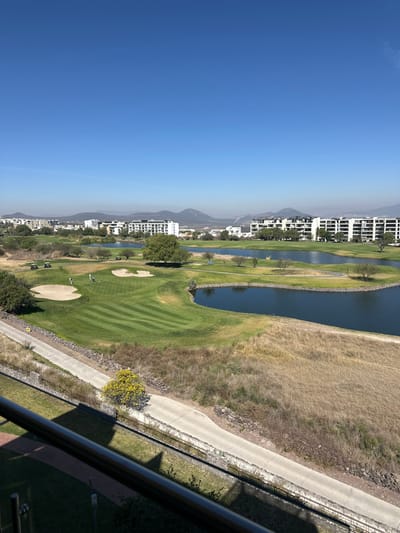 A serene, elevated view captures a golf course winding alongside a water feature, bordered by buildings against a hazy mountain backdrop. The manicured green contrasts with the adjacent terrain, divided by a paved path. A flowering tree dots the landscape, while an architectural detail partially obscures the lower right, hinting at the viewpoint.