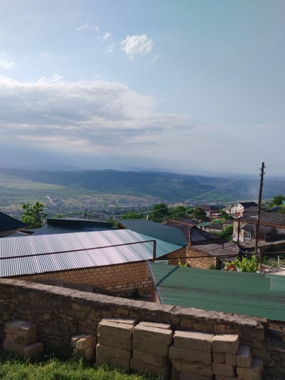 The image displays a panoramic view of Darvag, Russia, from an elevated vantage point. In the foreground, a low stone wall runs horizontally across the scene, with stacks of light-colored rectangular blocks resting against it on the right. Beyond the wall, several rooftops are visible, including corrugated metal roofs in silver and dark green, as well as some with a more weathered, darker appearance. The houses appear to be constructed with brick and stone.
Further into the background, the landscape descends into a valley dotted with more houses and buildings, forming a village or small town. This settlement is nestled amidst rolling green hills that stretch into the distance, gradually becoming more muted and hazy due to atmospheric perspective. The mountains in the far background are a deep blue-gray. A single wooden utility pole with wires extends vertically near the right edge of the frame.
The sky above is a mix of pale blue with scattered clouds. Some clouds are wispy and white, while others are larger and more textured, suggesting a partly cloudy day. The overall lighting indicates daytime, with sunlight illuminating the scene and casting shadows. The environment appears to be rural or semi-rural, characterized by natural topography and a collection of human dwellings.