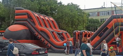 The image shows an outdoor scene featuring a large inflatable obstacle course with orange and black sections. Several people, including children and adults, are present, interacting with the inflatable structure. On the right side, a young boy is being suspended by a bungee harness attached to the inflatable. Other individuals are standing around, observing or participating.
In the background, lush green trees form a dense backdrop, suggesting a natural or park-like setting. Behind the trees, a light-colored building with multiple windows is visible, possibly a school or public facility. The sky is overcast, indicating a cloudy day. The overall atmosphere appears to be one of recreational activity, with families enjoying themselves.
To the left of the inflatable, several parked cars are visible, including a black BMW with a license plate that reads "C352BX 774". A woman wearing a blue sweater and a white backpack is walking away from the camera, passing by the cars. Another woman with an orange hat is standing near the inflatable.
The scene is likely taking place during the daytime, given the ambient light. The presence of the inflatable structure and the activities of the people suggest an event or a public playground setup. The setting seems to be a paved area, possibly a parking lot or an open space near a recreational facility. There are no strong indicators of specific emotions beyond general engagement and enjoyment of the outdoor activities.