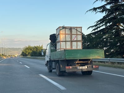 The image shows a green flatbed truck with its cargo bed filled with wooden crates, some of which are stacked on top of each other. The truck is traveling on a highway, with a clear blue sky overhead. There are trees and rolling hills in the background. The license plate of the truck is visible and reads "RR-437-YY" with a Georgian flag emblem. The truck appears to be in motion on the road. The scene is set in the vicinity of Dzveli Kanda, Georgia.