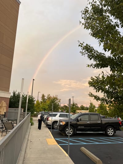 The image displays an outdoor scene in Tallahassee, United States, characterized by a prominent rainbow arcing across a pale, partly cloudy sky that exhibits faint orange and pink hues towards the horizon on the right. A multi-story, light brown building with horizontal paneling occupies the far left, featuring a brick base and an adjacent outdoor patio area equipped with at least two round tables, chairs, and a large beige umbrella, all enclosed by a metal railing. A concrete sidewalk with yellow tactile paving strips runs horizontally across the mid-ground. A single individual, observed from behind, walks on this sidewalk towards the right, wearing dark trousers, a light-colored top, and a dark backpack. To the right of the sidewalk lies an asphalt parking lot. A black Nissan Titan V8 crew cab pickup truck, distinguished by chrome accents and a tonneau cover, is parked in the foreground, with a silver SUV (possibly a Cadillac XT6) parked directly behind it. Additional vehicles are partially visible further in the lot. Green foliage from trees is scattered throughout the background and on the right side of the frame, partially obscuring distant structures. The natural lighting suggests late afternoon or early evening conditions.