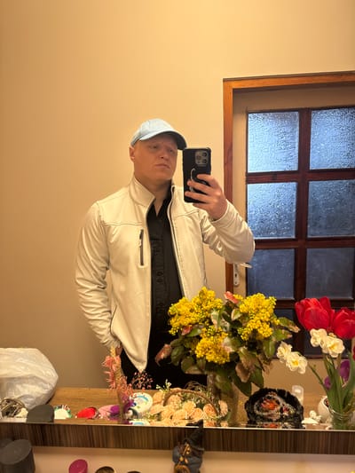 The image, currently rotated 90 degrees counter-clockwise, depicts an indoor mirror selfie. An adult individual, likely male, is shown from the chest up, holding a dark smartphone (likely an iPhone, visible through its multi-lens camera array) in their right hand to capture the reflection. The phone case features a white serpent or dragon design and legible white text that appears to read "WOOLRICH" and "STORY."
The person wears a light-colored, possibly off-white, collared jacket over a black collared shirt, and a light blue baseball cap. Their expression is neutral.
The background, reflected in the mirror, features a warm beige wall. A wooden-framed window with textured, frosted glass panes is visible, allowing diffused light from outside to enter the space, suggesting it is daytime, possibly overcast. To the right of the person, on a wooden surface, there are multiple floral arrangements, including a prominent bouquet of yellow mimosa-like flowers, red tulips, white narcissus-like blooms, and some purple flowers. Various other items are visible on the surface, including a black beaded accessory, several small decorative objects, hair clips, and a white plastic bag. The setting appears to be a domestic interior, possibly in Kavarna, Bulgaria.