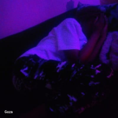 The image depicts an indoor scene, stated to be located in Kamera ward, Kenya, characterized by low light and a dominant purple hue. An individual is seated or crouched on a dark surface, possibly a sofa or bed, positioned in profile facing the left side of the frame. The person wears a white short-sleeved top and dark-colored trousers with a light-colored, abstract pattern. Both hands are raised to cover their face, obscuring facial features. A lighter, possibly textured, object is partially visible to the right of the individual, appearing to be a pillow or blanket. The background wall exhibits the same purple illumination. The text "Gaza" is visible in the lower-left corner of the image.
