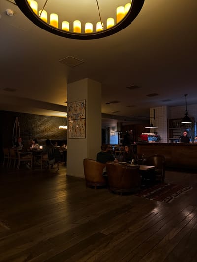 This is a low-angle, wide shot of the interior of a dimly lit bar or restaurant. The scene is captured in portrait orientation, with the camera looking upwards. Dominating the left side of the frame is a large, circular chandelier with multiple yellow lights. The rest of the interior features warm, wooden floors, exposed brick walls in the background, and dark, leather-upholstered chairs.
Several people are visible in the background, appearing to be patrons and staff. In the far background, a bar area is visible with people standing behind it. Some individuals are seated at tables or in chairs within the seating area. The overall atmosphere appears relaxed and cozy, with a focus on intimate lighting. The time of day is likely evening or night, given the interior lighting and the dark ambiance. There is no indication of the weather outside.
On a wall in the mid-ground, a framed poster or artwork is visible with some text. The text appears to be in English, with "EUROPEAN PHOTOGRAPHY" and "WEIBIG" being partially legible. This poster might suggest the venue has an artistic or cultural theme. The setting appears to be a modern establishment designed for social gathering and relaxation.