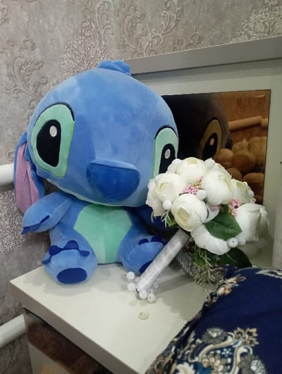 A blue Stitch plush toy rests on a table with a white bouquet of flowers. The bouquet features white roses and small pink flowers, adorned with tiny white pearls. The plush toy is positioned on the left side of the frame, while the flowers occupy the upper right corner. The table is white with a reflective edge. In the background is a framed picture and wallpaper.