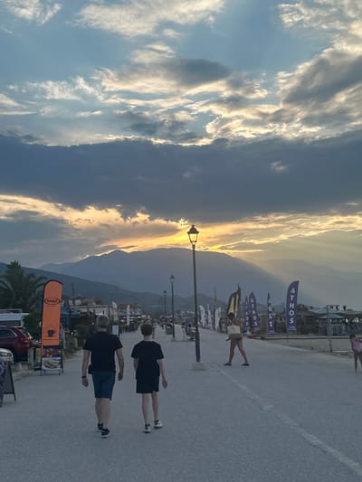 This media file captures a scene in Νέοι Πόροι, Greece, during what appears to be late afternoon or early evening. The sky is dominated by dramatic clouds, with streaks of golden sunlight breaking through, suggesting a sunset or a similar atmospheric phenomenon. In the background, mountains are visible, partially obscured by the low-lying clouds and the light.
The foreground shows a paved area, possibly a promenade or street, with several people. Two individuals in the foreground are walking away from the camera, both wearing dark t-shirts and shorts. Further down the paved area, other people are scattered, some appearing to be engaged in activities, while others are standing or walking. A woman in a pink top and white shorts is visible in the distance, on the right side of the frame.
Along the edge of the paved area, there are several blue flags with white text, the most prominent reading "ATHOS". A lamppost stands near the center of the image, illuminated. Various structures, possibly buildings or stalls, line the right side of the scene, with some appearing to be shops or cafes. An orange banner with text in a foreign language is visible towards the bottom of the frame, and a palm tree is situated near it. The overall atmosphere is relaxed and summery, with the natural beauty of the sky and landscape contrasting with the human activity.
