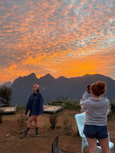 Two women stand near a mountain range at sunset. One woman, with red hair, sits on a white plastic chair and takes a photo with her smartphone. She wears a gray hoodie and dark blue shorts. The other woman, wearing a colorful headscarf and denim jacket, stands on a dirt surface. A metal and wood platform with railing is next to her. The sky displays a vast, orange-tinted cloud formation.