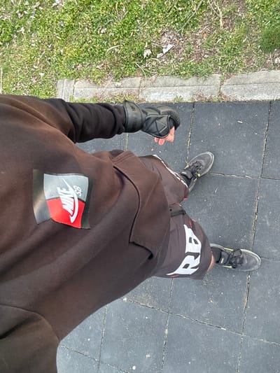 The image, taken in Dolno Sahrane, Bulgaria, provides a high-angle view of a subject standing on a dark grey, square-tiled rubberized surface. The subject wears a dark brown long-sleeved top with a prominent, partially detached Nike SB logo patch, rendered in grey, black, and red, on the back or side. A black, padded protective glove, with "RITUAL" text visible on its wrist strap, is worn on the subject's left hand. The subject is also clad in dark athletic shorts or trousers, which display white "RBA" lettering on the right thigh. Dark athletic shoes with light accents on the soles are visible on the subject's feet, positioned on the tiled surface. Immediately adjacent to the rubberized area is a concrete curb, beyond which lies an expanse of green grass containing sparse brown debris. The scene is illuminated by natural light.