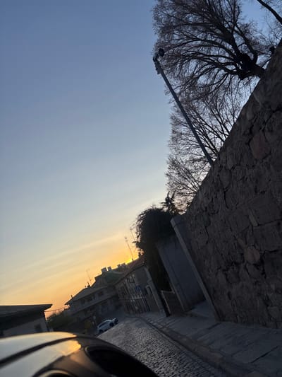 The image captures a tilted, low-light street scene in the city of Ávila, Spain, during either sunrise or sunset, evidenced by the orange-yellow horizon. The sky is clear, transitioning from blue overhead to yellow-orange near the horizon. A robust, irregularly coursed stone wall, composed of dark grey and brown blocks, dominates the right side of the frame, angling upward and right. Beyond and above this wall, the skeletal branches of bare deciduous trees are silhouetted against the sky, along with a tall, grey metal streetlight pole. The street itself is paved with cobblestones, slopes downhill from right to left, and curves subtly. On the left side of the street, multiple multi-story buildings with varied architecture and rooftop antennas are visible. A white compact SUV or crossover vehicle is parked on the left side of the cobbled street, facing uphill. A blue and white road sign is discernible on the left sidewalk further down the street. The bottom left foreground shows a dark, reflective surface, likely part of a vehicle's dashboard or frame, reflecting the sky's orange glow. No people are present, and no active events are depicted.