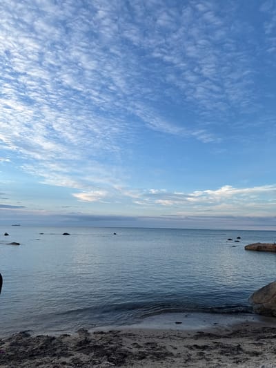 The image depicts a serene seascape with a calm ocean meeting a sandy beach in Rohuneeme, Estonia. The sky above is a vibrant blue, dotted with scattered, wispy white clouds that create a textured, patterned effect. The horizon is visible, separating the sea from the sky, and in the distance, a large ship can be seen sailing on the water. Several large rocks emerge from the tranquil sea, breaking the otherwise unbroken expanse of water. The beach in the foreground consists of dark sand mixed with small pebbles and debris, with gentle waves lapping at the shore. The overall atmosphere is peaceful and natural, characteristic of a coastal landscape.