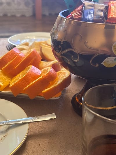The image shows a table setting at what appears to be a home or cafe in Getk, Armenia. In the foreground, a glass with a dark liquid, likely a beverage, is visible. To the left, a partially eaten plate of sliced oranges and what seem to be apple slices is presented. A fork rests on the plate. In the background, a decorative bowl filled with wrapped candies is placed on a stand. The overall scene suggests a moment of rest or a small gathering.