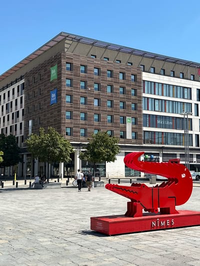 The image shows a street scene in Nîmes, France, under a clear blue sky, indicating it is daytime. In the foreground, a large, stylized red sculpture is prominently displayed. The sculpture is a metallic, layered representation of what appears to be a crocodile head or a similar reptilian creature, with sharp, jagged edges. On the side of the sculpture's base, the word "NÎMES" is clearly visible in white lettering.

Behind the sculpture, a modern, multi-story building with a distinct architectural style dominates the background. The building's facade features a pattern of horizontal wooden slats interspersed with rows of windows. Several flags or banners are visible on the building, including green banners with the "ibis styles" logo and blue banners with the "ibis budget" logo, suggesting the presence of these hotel brands. Higher up on the building, text such as "Midi Libre" can be read on some of the windows, indicating a media company.

Several people are scattered throughout the scene, mostly on the paved plaza in front of the building. Some are walking, while others are sitting or standing, engaged in conversations. One group near the sculpture appears to be a mix of adults and possibly children, some with a bicycle. The overall atmosphere seems relaxed, with people enjoying the outdoor space. The sunlight casts shadows on the plaza, further confirming it is a bright, sunny day.