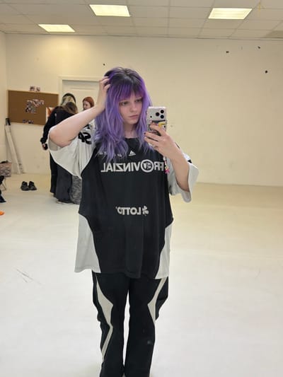 In an indoor room in Lubnya, Russia, a young individual with long, wavy purple hair and bangs stands facing a mirror, holding a smartphone in their right hand to capture a selfie. Their left hand is raised, fingers touching the hair on the left side of their head. The individual wears an oversized black and white athletic-style t-shirt, which appears to have a mirrored and inverted "PROVINCIAL" logo and a "LOTTO" logo, along with black pants featuring white stripes along the outer seams. The room features light-colored walls and a matching floor, with square fluorescent light fixtures recessed into the ceiling. In the reflected background, behind the main subject, a bulletin board with various papers is visible on the left wall, near a white door. The reflections of at least two other individuals can be seen: one with lighter hair to the left and another with reddish hair further back. A pair of dark shoes and a small container are visible on the floor in the reflected background. The right side of the reflected background consists of a plain, light-colored wall.