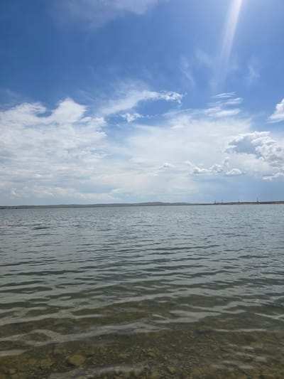 A wide, tranquil body of water stretches across the frame, its surface rippling gently under a partly cloudy sky. The water is clear enough in the foreground to reveal a rocky, shallow bottom, giving way to deeper, darker hues further out. The horizon is marked by a low, undulating line of hills under a vast expanse of blue sky dotted with white and wispy clouds. A bright sunbeam pierces through the clouds in the upper right corner, casting a luminous glow. Along the distant shoreline, a series of tall, slender poles with crossbars are visible, suggesting some form of infrastructure, possibly related to power transmission or a communication tower. The scene evokes a sense of open space and natural beauty, with a hint of human presence on the distant shore. The overall impression is one of a peaceful, expansive natural landscape.