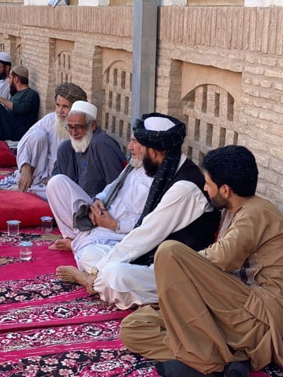 In the city of Kandahar, Afghanistan, a group of men are seated on a patterned red carpet, a common setting for gatherings. The men, dressed in traditional clothing, are facing each other, suggesting a social or religious occasion. The background features a brick wall with decorative arched recesses. The lighting suggests it is daytime, possibly late afternoon or early evening, given the shadows.