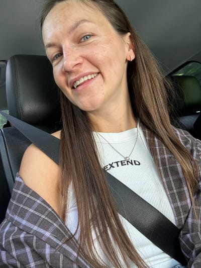In the city of Miravčine, Montenegro, a young Caucasian woman with long brown hair and blue eyes is seated in the passenger seat of a vehicle, wearing a white ribbed tank top with the word "EXTEND" printed on it in black letters, and a plaid shirt draped over her shoulders. She is smiling and looking directly at the camera, with a seatbelt fastened across her chest. The interior of the car is visible, including a black leather seat.