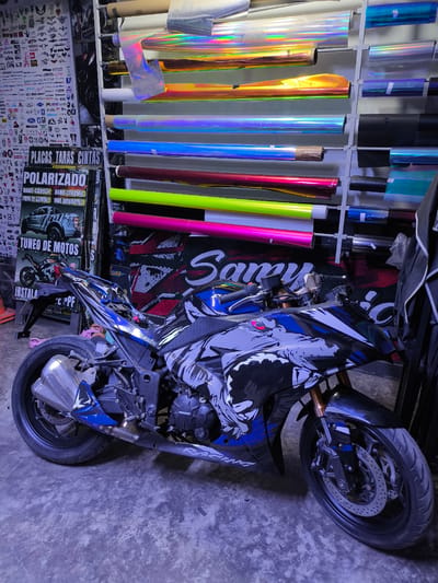 The image shows a motorcycle in what appears to be an auto customization shop. The motorcycle is blue and black with a wolf graphic. Rolls of brightly colored vinyl are displayed on shelves in the background, along with posters for car and motorcycle accessories. A sign with "POLARIZADO" and "TUNEO DE MOTOS" indicates services offered, such as window tinting and motorcycle customization. The sticker wall also suggest this is a tuning shop, and the "Samu" logo in the background also appears to refer to auto customization. "Trujillo" and "Peru" are not mentioned in the OCR text or discernible from the scene.