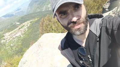 A selfie shows a man with a beard and a cap, dressed in a dark jacket and t-shirt, standing on a rock overlooking a landscape. In the background, a valley with a town is visible, along with mountains. The location is Sini Vrah, Bulgaria. The man is looking at the camera with a slight smile. Sunglasses hang on his shirt.