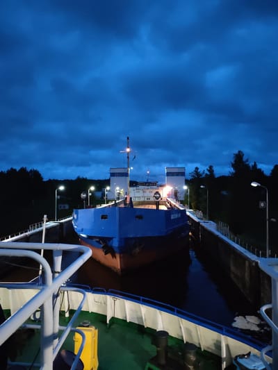 In the city of Devyatiny, Russia, a blue and red cargo ship is docked in a canal lock. The sky is a deep, cloudy blue, indicating dusk or night. The lock walls are made of concrete, with metal railings and lampposts lining the sides. The ship's name, "ОКСКИЙ-59," is visible on its hull. The foreground shows part of another vessel, with green decks and white railings.