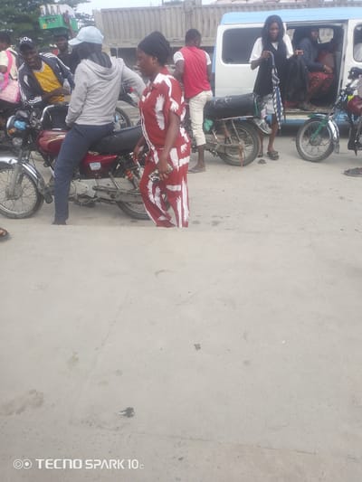 In Adesanya, Nigeria, a group of people are gathered around motorcycles and a van. A woman in a red and white patterned dress walks towards the viewer. Behind her, a man wearing a grey hoodie and a light blue cap sits on a motorcycle. Another man, wearing a yellow shirt and a black jacket, also sits on a motorcycle to the left, with a bottle of water in his hand. To the right, a man in a red t-shirt and white pants stands near a motorcycle, while a woman in a white shirt and black vest stands with bags by an open van. The scene appears to be a busy street or transport hub, with a truck visible in the background. The ground is paved and appears dusty.