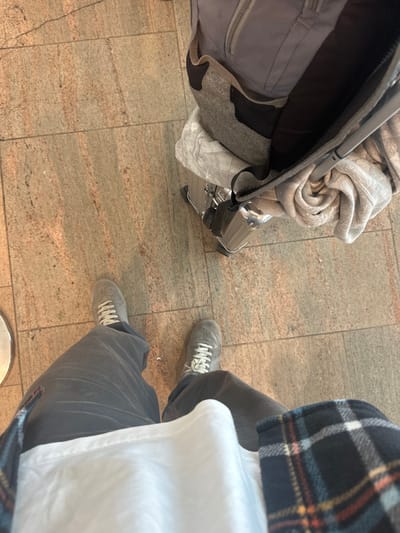 A first-person, downward-facing view captures the lower half of an individual standing on a light brown tiled floor within what appears to be an airport terminal in Israel. The person wears dark gray pants, a white t-shirt, and a dark blue, plaid flannel shirt with accents of orange, light blue, and white. Gray athletic shoes with white laces are worn on both feet. To the individual's right, a gray backpack with black mesh side pockets and shoulder straps rests partially on a silver-colored, ribbed hard-shell suitcase. A light gray, rolled-up garment is partially visible, tucked into the top opening of the suitcase. The floor consists of rectangular tiles with visible grout lines and subtle color variations.