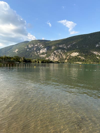 This media file depicts a serene lakeside scene in Novalaise, France, likely during a warm, sunny day. The image is oriented vertically, with the water occupying the right side and the sky and a mountain on the left.
The foreground features clear, shallow water with a sandy or pebbly bottom visible through the ripples. The sunlight creates a shimmering effect on the water's surface and casts intricate patterns on the submerged ground. Along the water's edge, a series of wooden posts are submerged, with a white rope strung between them, suggesting a marked swimming area or a boundary. A few yellow buoys are also visible in the water, further indicating a designated zone.
The background is dominated by a lush, green mountain that slopes down towards the lake. The mountain is densely covered with trees, with rocky outcrops visible in places. The sky above is a vibrant blue with scattered white clouds, some of which are fluffy and bright, while others appear more wispy and elongated.
The overall mood of the image is peaceful and tranquil. The clear water, the verdant mountain, and the bright sky evoke a sense of natural beauty and relaxation. The time of day appears to be late morning or early afternoon, given the bright sunlight and the clear sky. There are no visible people or specific events taking place, making it a snapshot of a quiet moment in nature. No text is visible in the image.