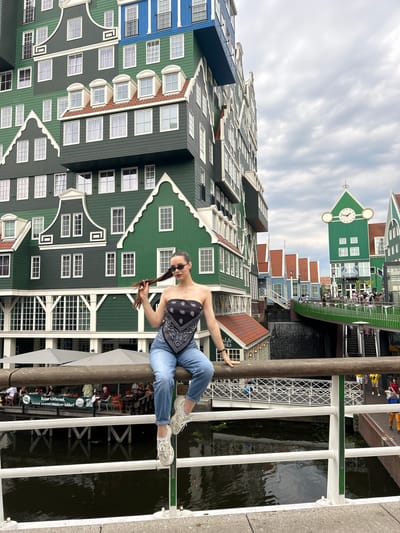 This is a vertical, eye-level shot of a young woman posing for a photo in front of a row of brightly colored, traditional Dutch houses in Zaandam, Netherlands. The woman is wearing blue jeans, a black bandana top, and white sneakers. She is sitting on a railing, with her legs extended, and holding her hair. She is wearing sunglasses and has a relaxed expression.
The houses behind her are a mix of green, blue, and red, with white trim and many windows. One of the buildings has a prominent clock face. The scene appears to be a canal or waterway, with a bridge or walkway in the foreground where the woman is sitting. There are other people visible in the background, some sitting at tables outside a cafe.
The weather appears to be overcast, with a cloudy sky. The lighting suggests it is daytime, possibly late morning or early afternoon. The architecture of the buildings strongly indicates a Dutch location, and the provided context confirms it is Zaandam. There is text on a green banner visible in the background which reads: "Buiten schitterend. Binnen onweerstaanbaar lekker!".