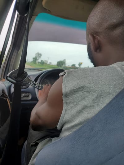 The image shows a person driving a vehicle, with a view from inside the car. The primary subject is the driver, whose head and upper body are visible. They are wearing a grey t-shirt and blue denim jeans. The driver's right arm is on the steering wheel, and their hand is partially visible. A seatbelt is prominently displayed across the frame, secured at the buckle.
The scene is set inside the car, with the dashboard and steering wheel visible. Beyond the windshield, a slightly overcast sky and green trees suggest an outdoor environment, possibly a rural or suburban setting. The weather appears to be cloudy or hazy, indicating it could be morning or afternoon with less direct sunlight. The location context of Pankshin, Nigeria, suggests a road trip or daily commute within that region.
A notable detail is a monkey wrench attached to the seatbelt, which appears to be used as a makeshift way to hold the seatbelt in place or possibly to adjust it. This detail is unusual and might suggest a practical improvisation. There is no visible text in the image. The overall mood is functional, focused on the act of driving.