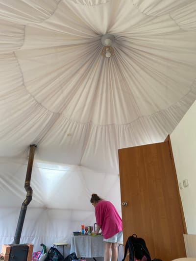 In a yurt-like structure, a woman in a bright pink shirt and light blue shorts stands bent over a table, seemingly preparing food or drinks. The interior is white and fabric-covered, with a central light fixture. A metal stovepipe extends from the floor upwards on the left, near a brick base. A wooden door is visible on the right. The environment appears to be a temporary or glamping accommodation. The image is captured from a low angle, looking up towards the tent's ceiling.