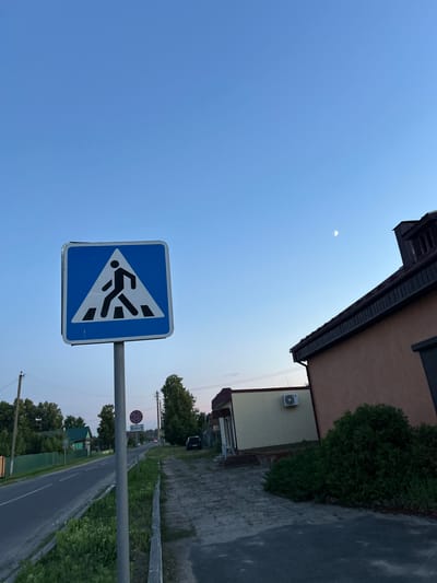 In the town of Parytsky rural Council, Belarus, a pedestrian crossing sign stands prominently on a metal pole on the side of a road. The sign, a blue square with a white triangle containing a black silhouette of a person walking, indicates a designated crosswalk. Beyond the sign, a paved sidewalk runs alongside the road, bordered by lush green grass and small bushes. A two-story building with a brown facade and dark roof is visible to the right of the sidewalk. The sky above is a clear, pale blue, with a crescent moon faintly visible. The road stretches into the distance, lined with trees and houses, suggesting a residential area. A secondary sign with a red circle and arrow, indicating a no-stopping zone, is also present on the pole, below the pedestrian crossing sign. The scene depicts a quiet, rural street in the late afternoon or early evening.